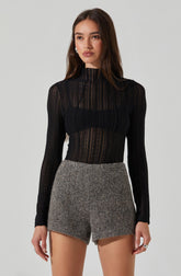 A young woman is wearing a black, long-sleeved, semi-sheer, high-neck top with vertical lace patterns. She pairs it with high-waisted, gray, textured shorts.
